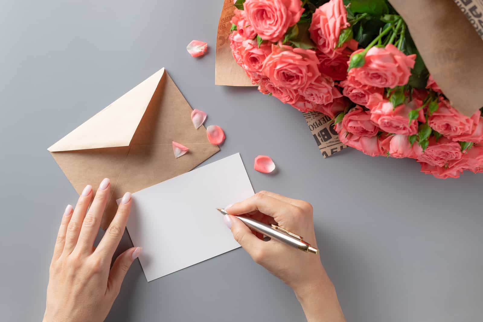 Frau, die Grußkarte auf grauem Tisch mit Strauß rosa Rosen schreibt