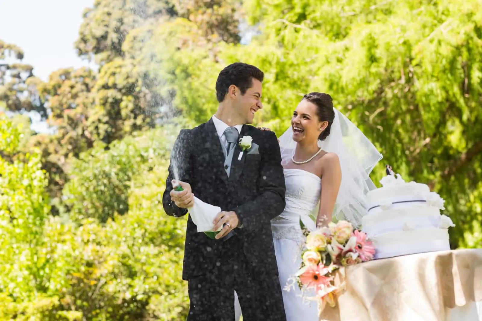 Glückliches junges frisch verheiratetes Paar mit Bräutigam, der die Champagnerflasche am Park öffnet