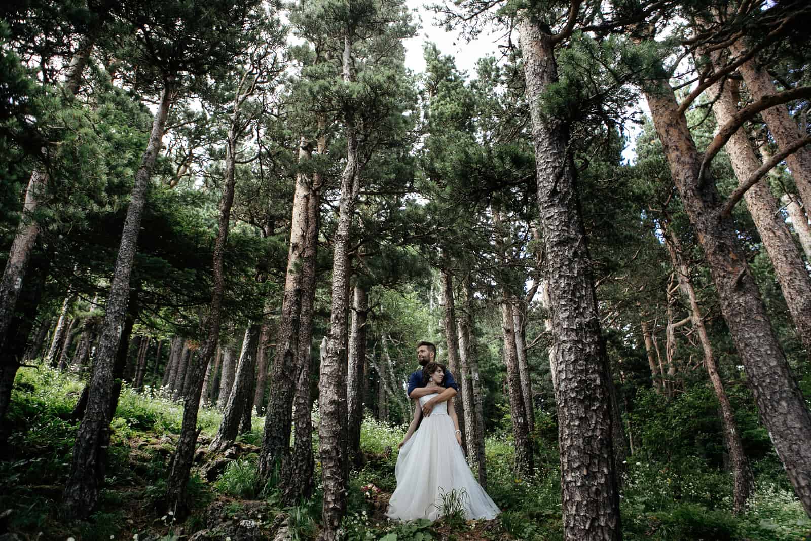 der Bräutigam mit Bart in einem blauen Hemd und die Braut auf einem Hochzeitsspaziergang im Wald in den Bergen