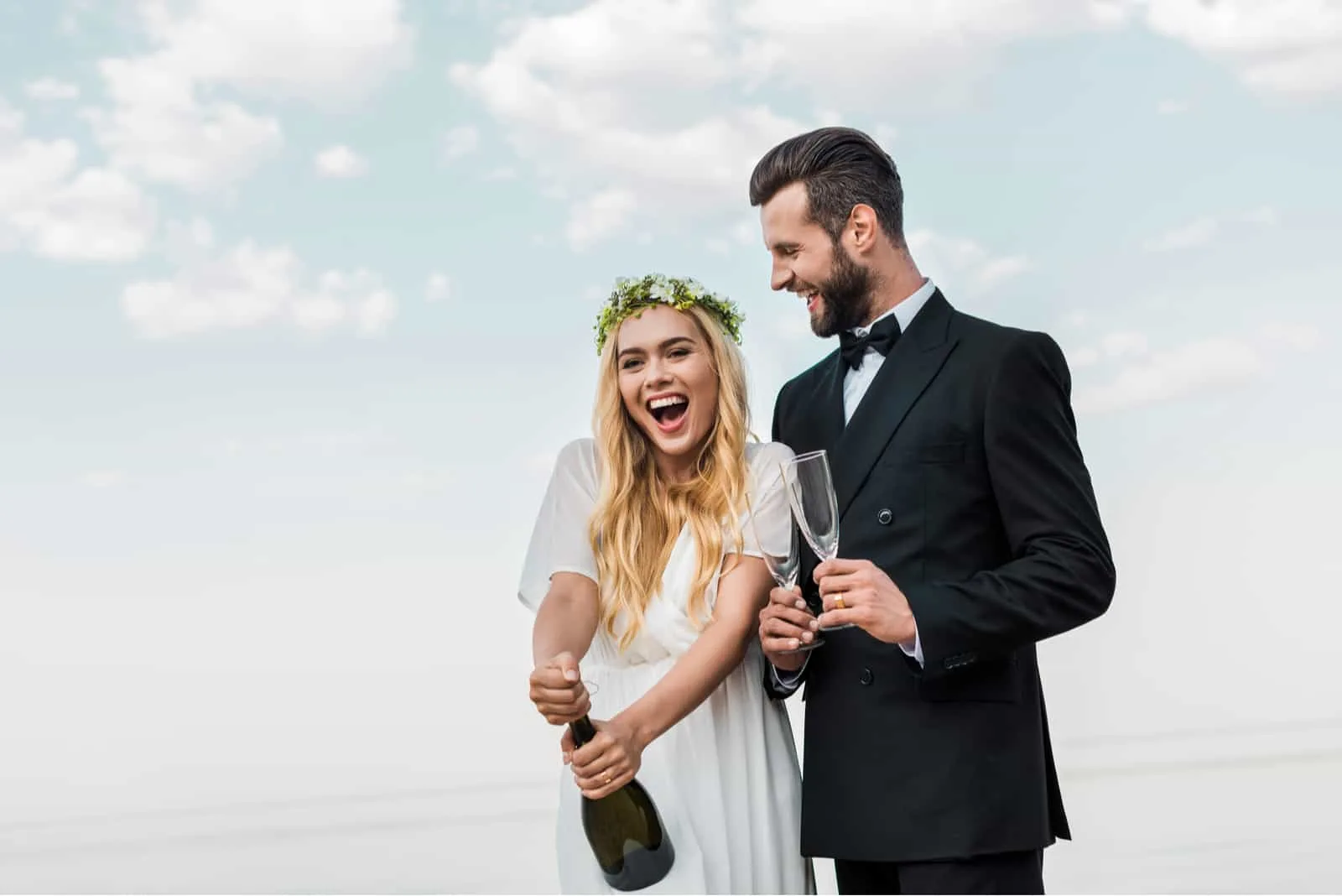 glückliche Braut im weißen Kleid, das Champagnerflasche am Strand öffnet