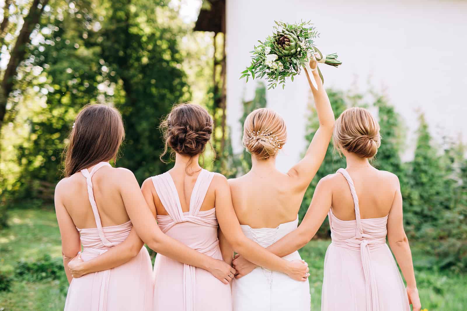 Braut, die mit dem Rücken in einem weißen Hochzeitskleid und erhabener Hand mit drei Brautjungfern steht