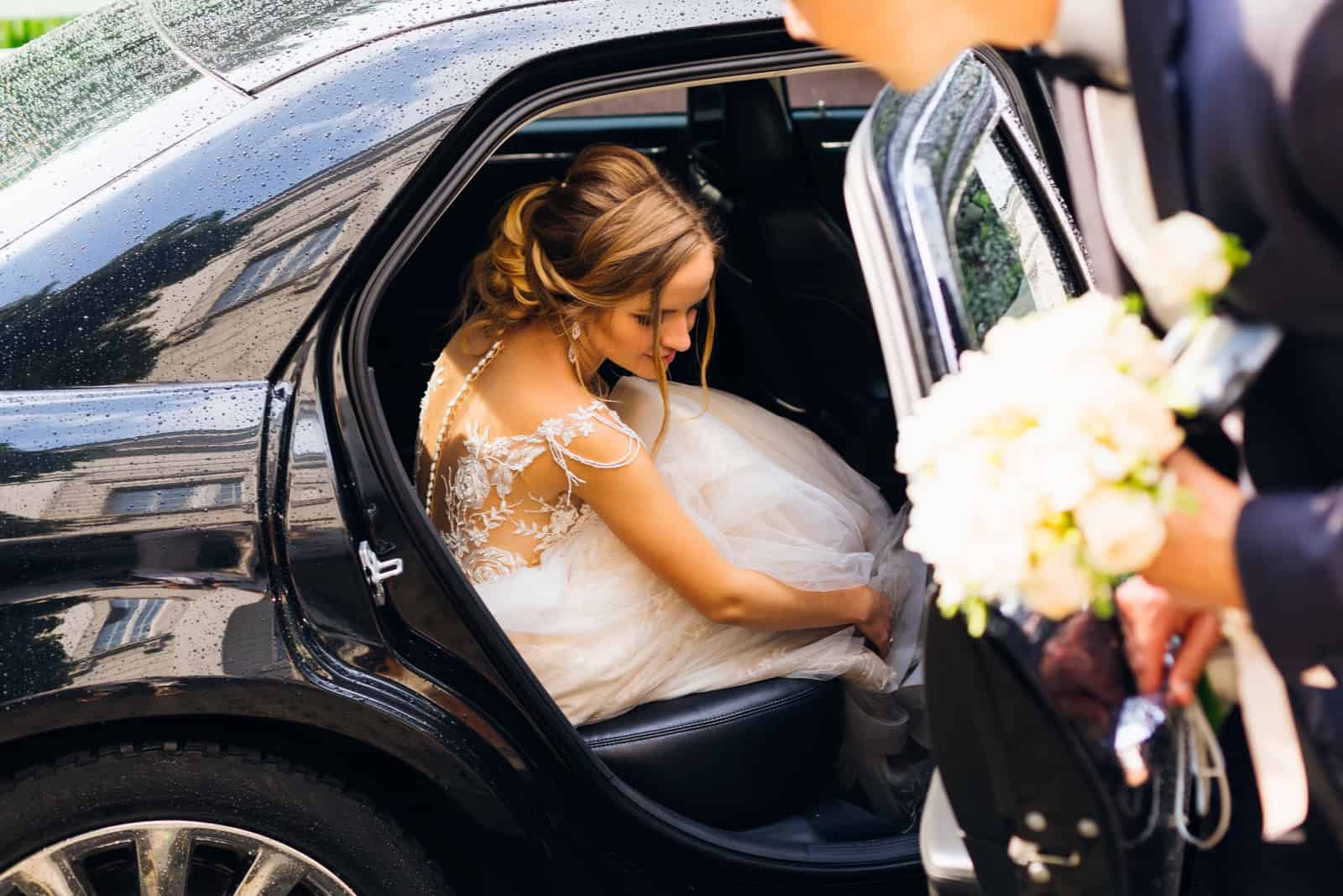Die schöne Braut sitzt in einem schwarzen Auto und der Bräutigam will die Tür danach schließen