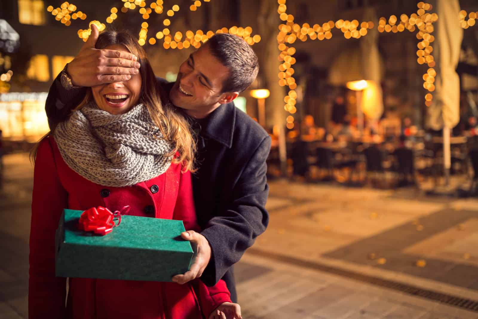 Mann hält die Augen seiner Freundin bedeckt, während sie ein Geschenk gibt