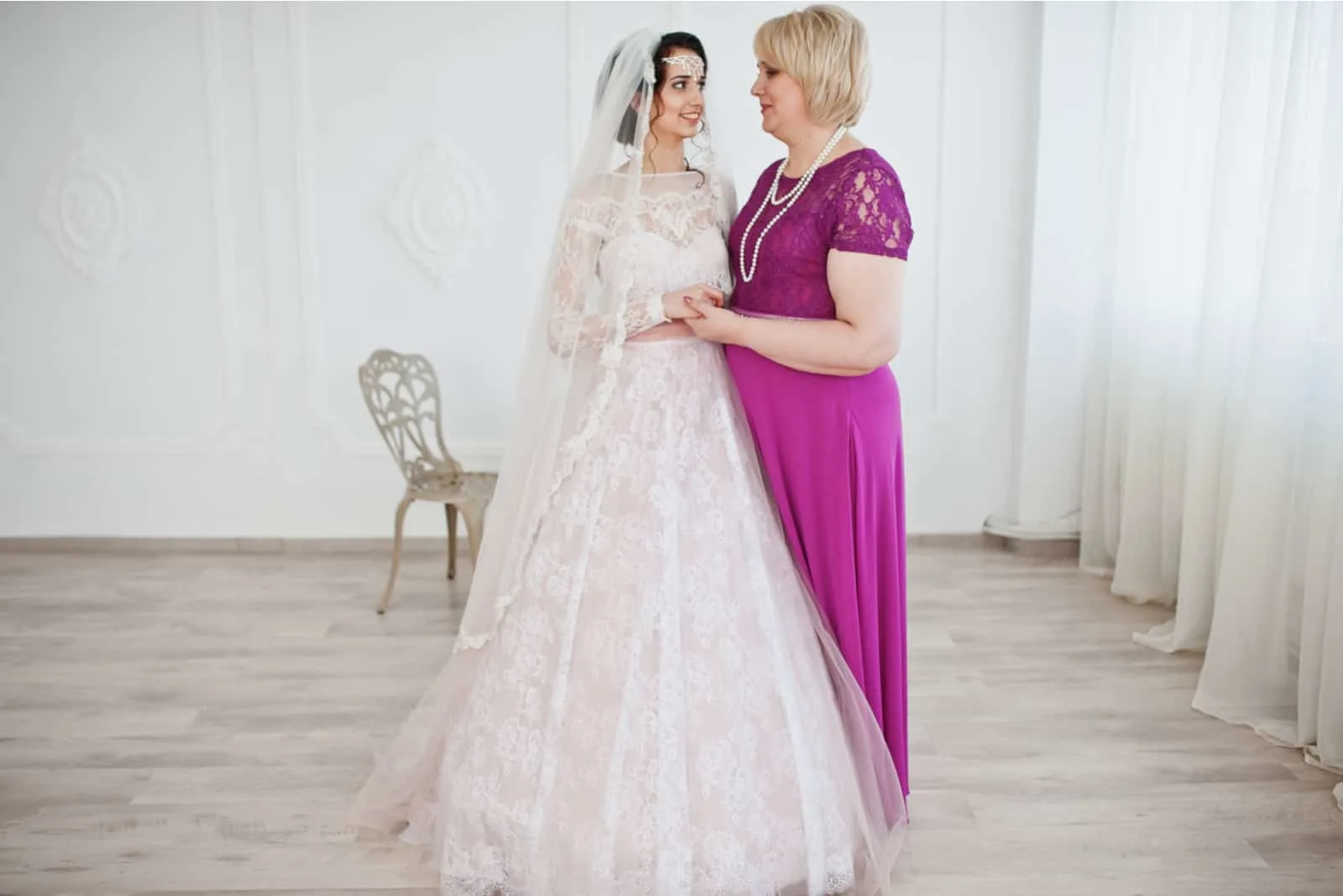Schöne Braut im Hochzeitskleid posiert mit ihrer Mutter im lila langen Kleid.