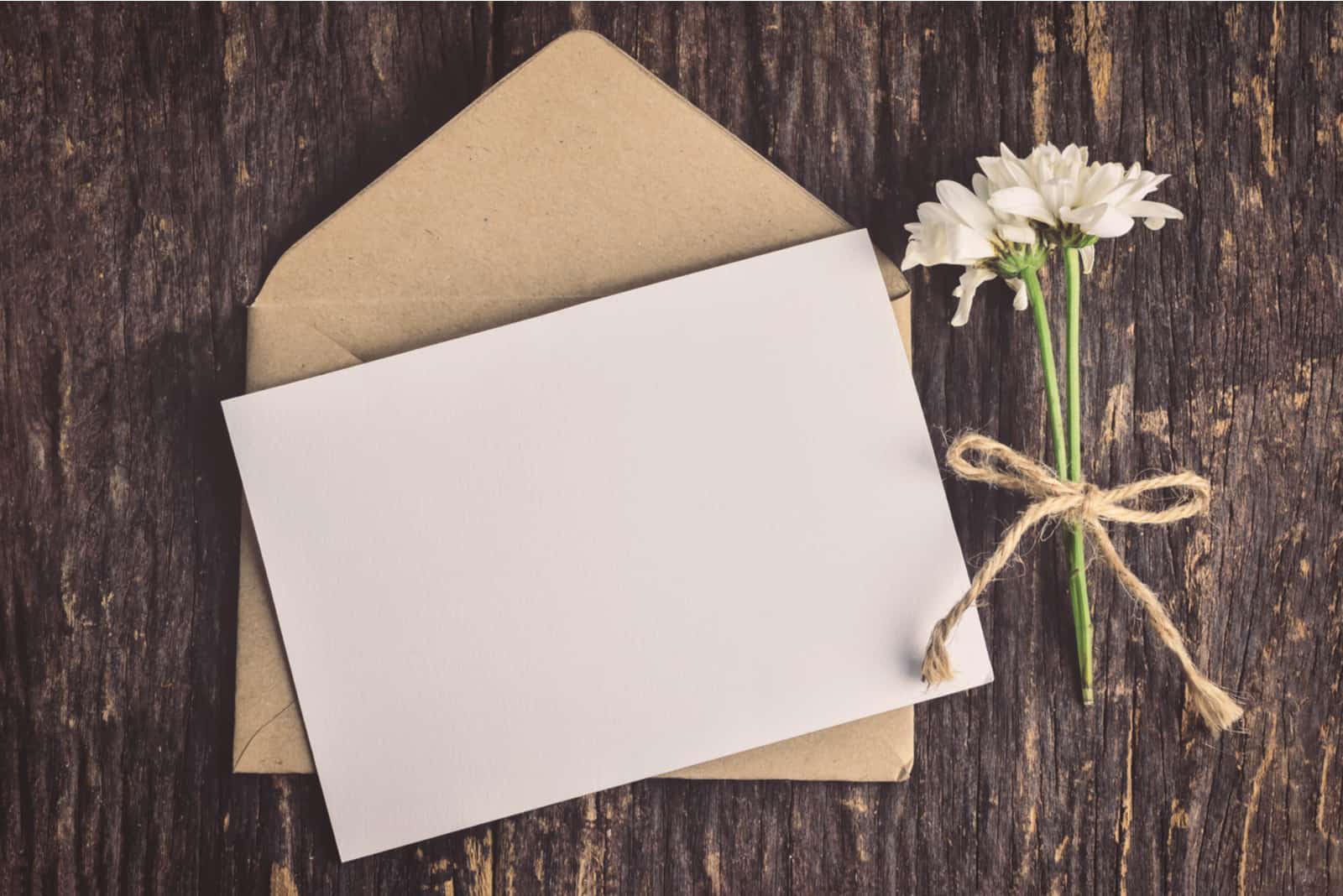 weiße Grußkarte mit braunem Umschlag und verwelkten Mutterblumen auf Holztisch