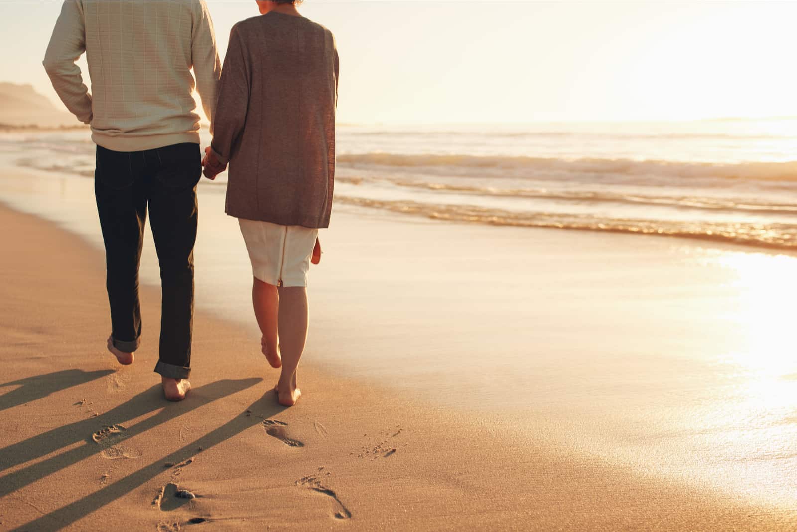 älteres Paar Händchen haltend und bei Sonnenuntergang am Strand spazieren gehen