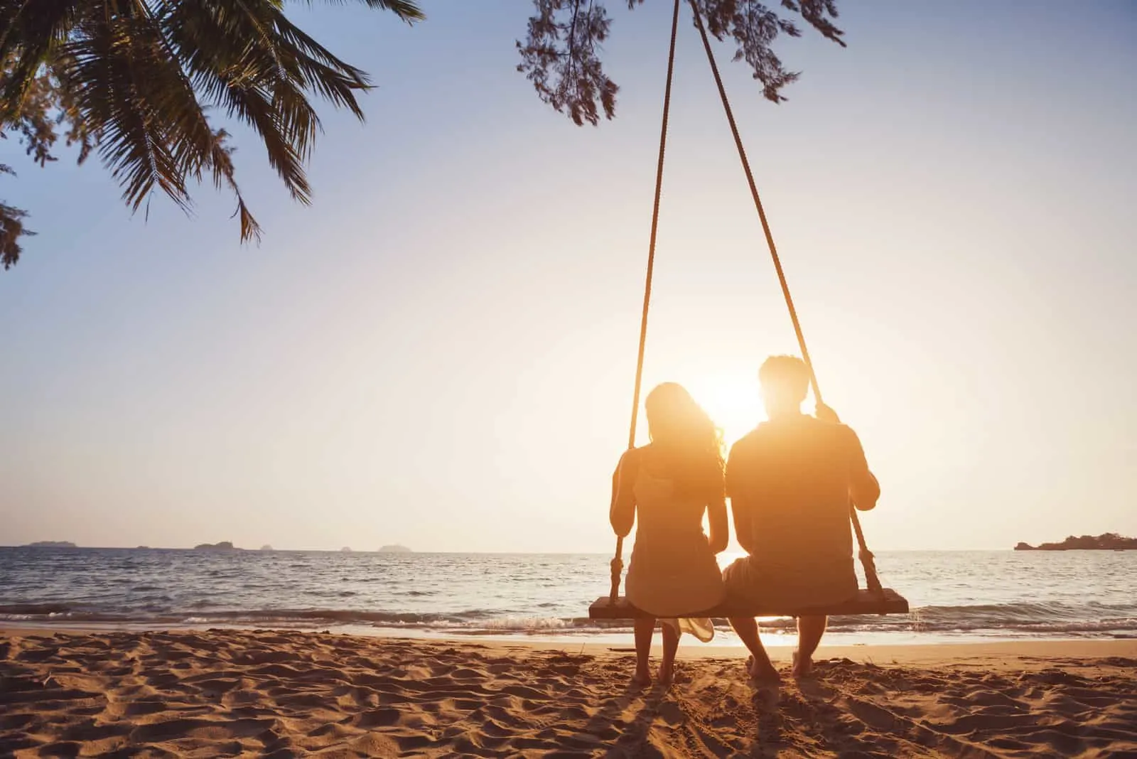 romantisches verliebtes Paar, das zusammen auf Seilschaukel am Sonnenuntergangstrand sitzt